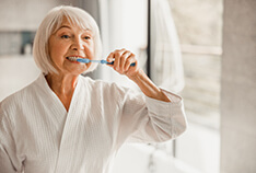 An older woman wearing a white robe brushes her teeth while in her bathroom
