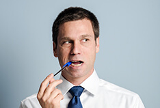 A man wearing a button-down shirt and blue tie bites down on the tip of a pen