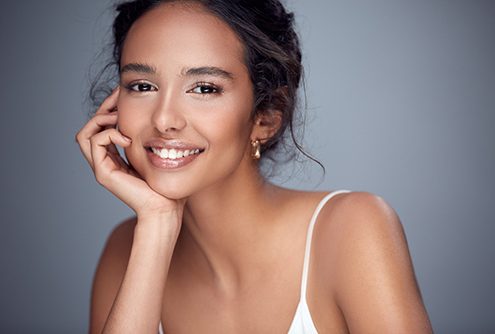 A young woman wearing a white tank top with her hair pulled back and resting her face in her propped hand and smiling after seeing a dentist about a smile makeover in McMinnville