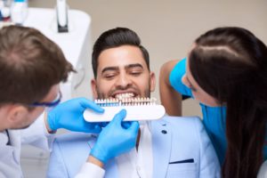 Dentist and associate hold a shade guide to the teeth of a man in a pale blue suit
